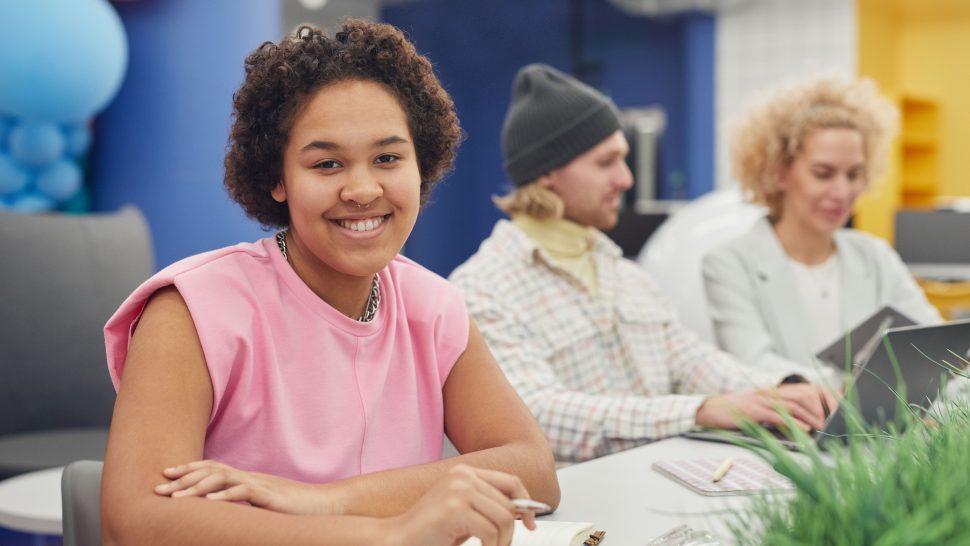Young woman sitting at a table with other young adults in the background.